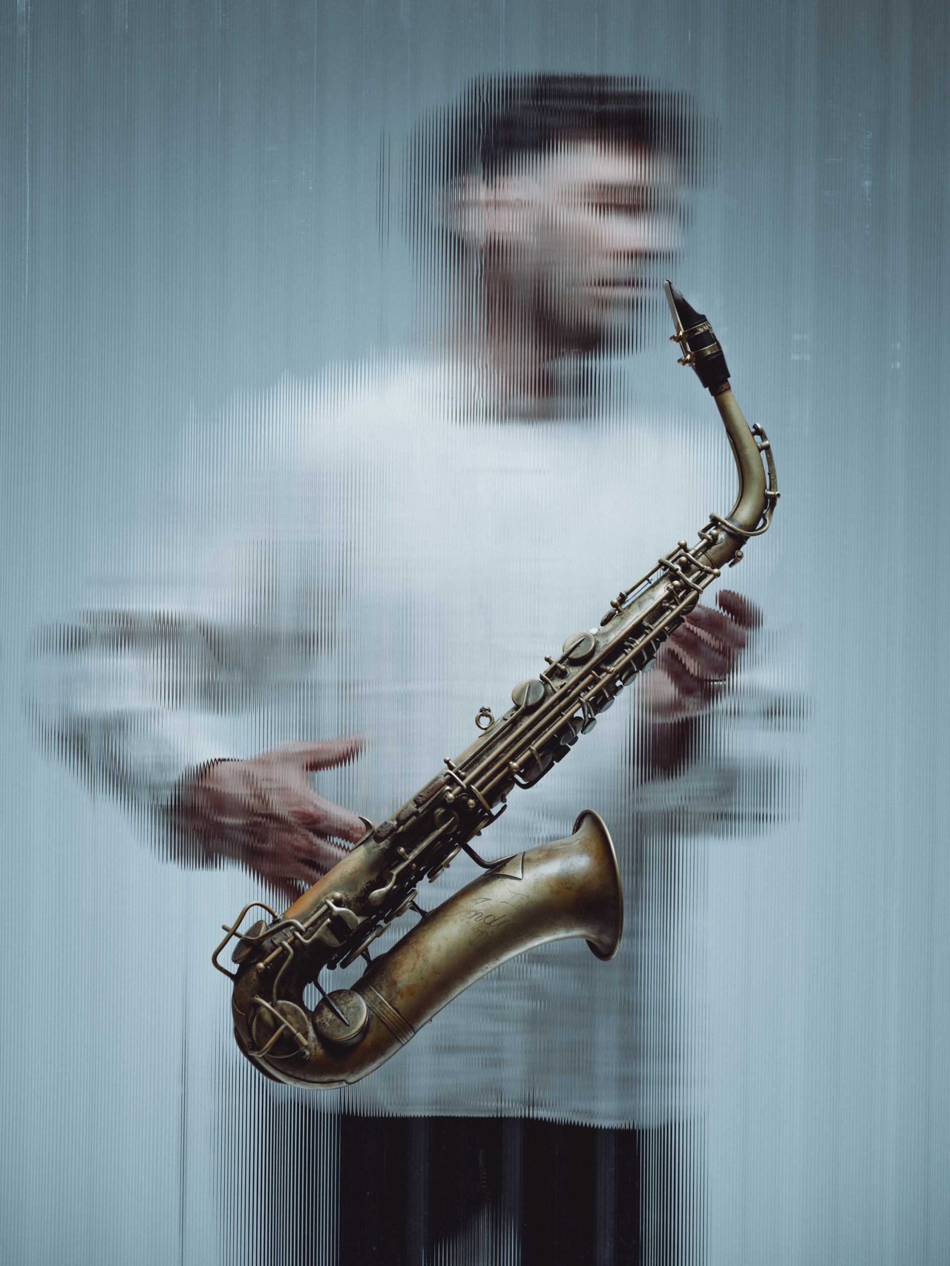 Künstlerisches Portrait des Musikers Christian Weidner mit seinem Saxofon. Der Musiker steht hinter einer Scheibe Riffelglas, das Instrument hängt vor der Glasscheibe.